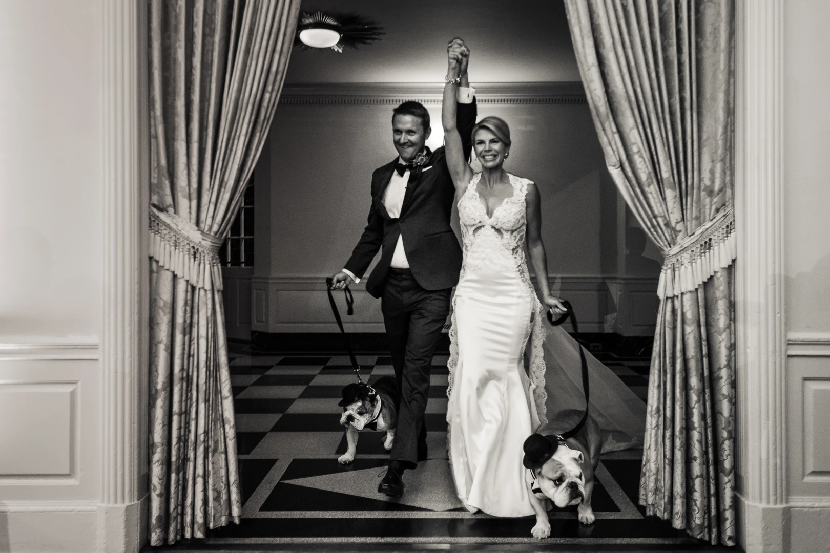 A joyful bride and groom walk through elegant curtains holding hands, each leading a dog on a leash. The setting is a formal, classic venue with checkered floors and ornate drapery. The bride wears a long gown, and the groom is in a suit.