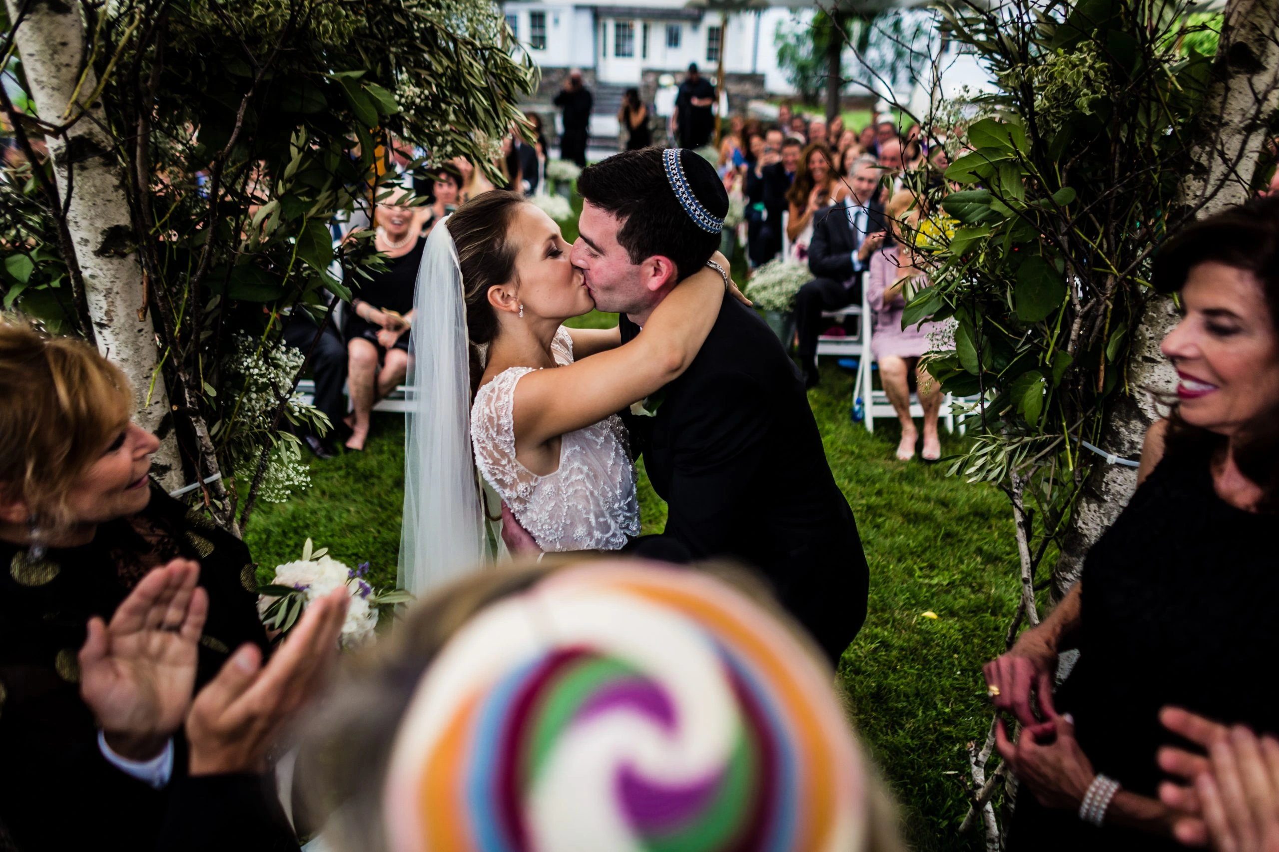 A bride and groom share a kiss under an archway of greenery at an outdoor wedding ceremony. Guests, seated in the background, are clapping and celebrating. The bride wears a veil, and the groom has a kippah on his head.