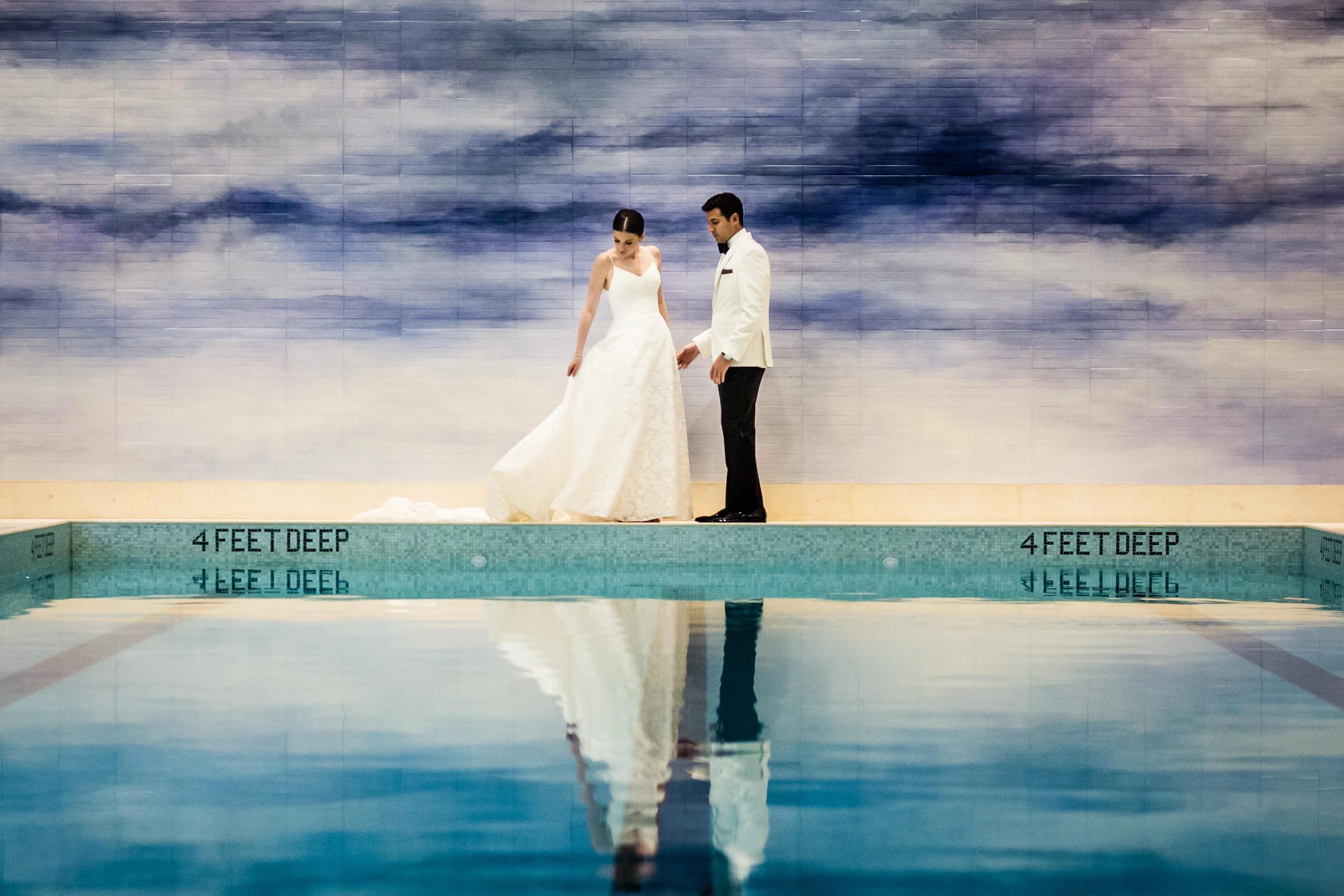 A bride and groom dressed in formal attire stand near the edge of an indoor pool, labeled "4 feet deep." The pool reflects their image against a blurred sky-like backdrop.