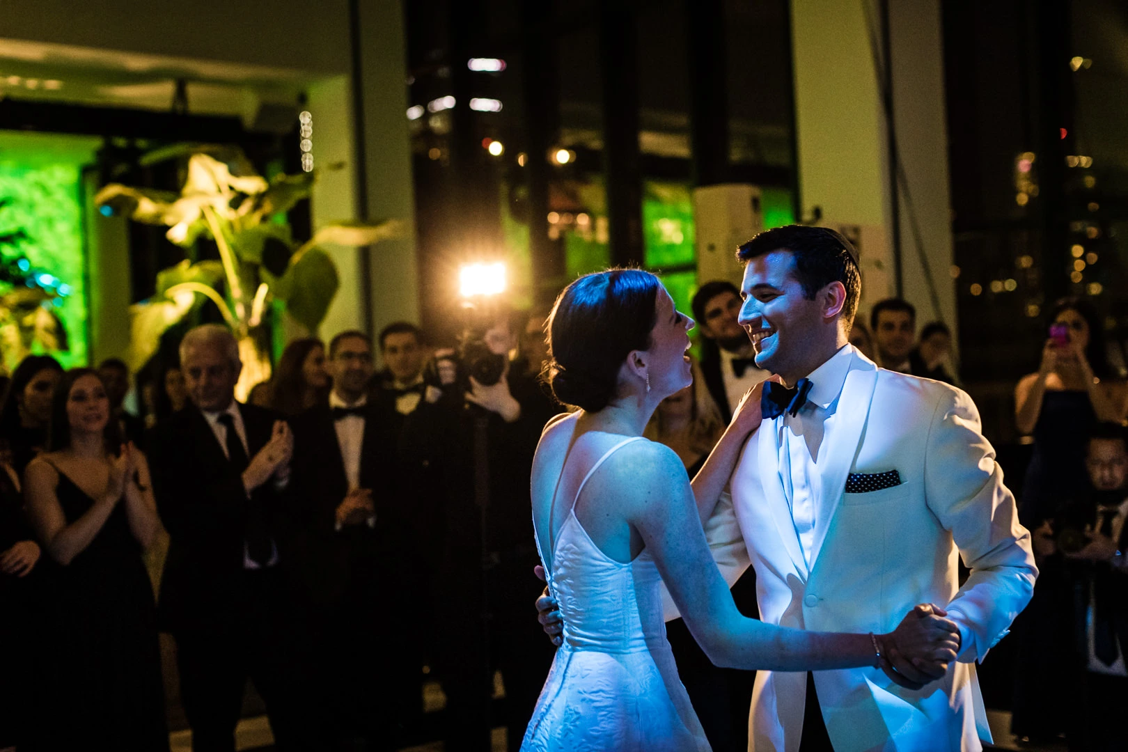 A bride and groom dance joyfully under colorful lighting at their wedding reception. Guests, dressed in formal attire, watch and smile in the background.