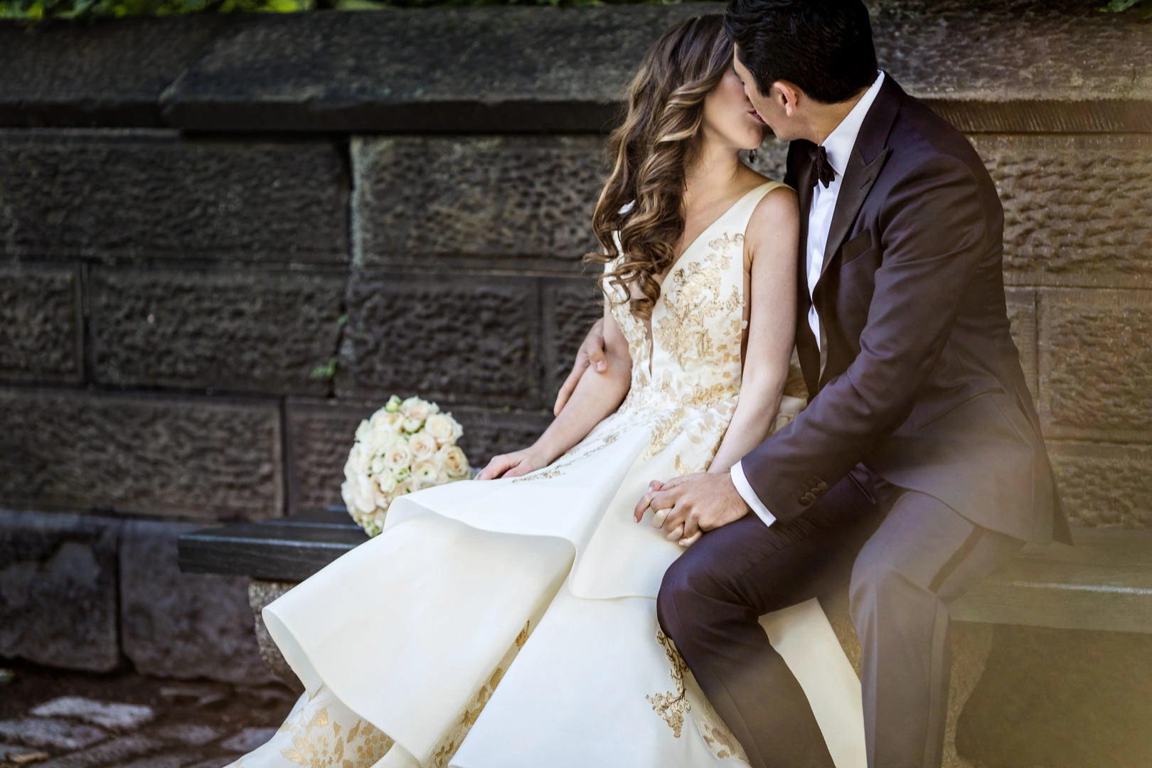 A couple in formal attire sits on a bench, sharing a kiss. The woman wears an elegant white and gold dress and holds a bouquet of roses. The man is in a dark suit with a bow tie. They are in an outdoor setting with a stone wall in the background.