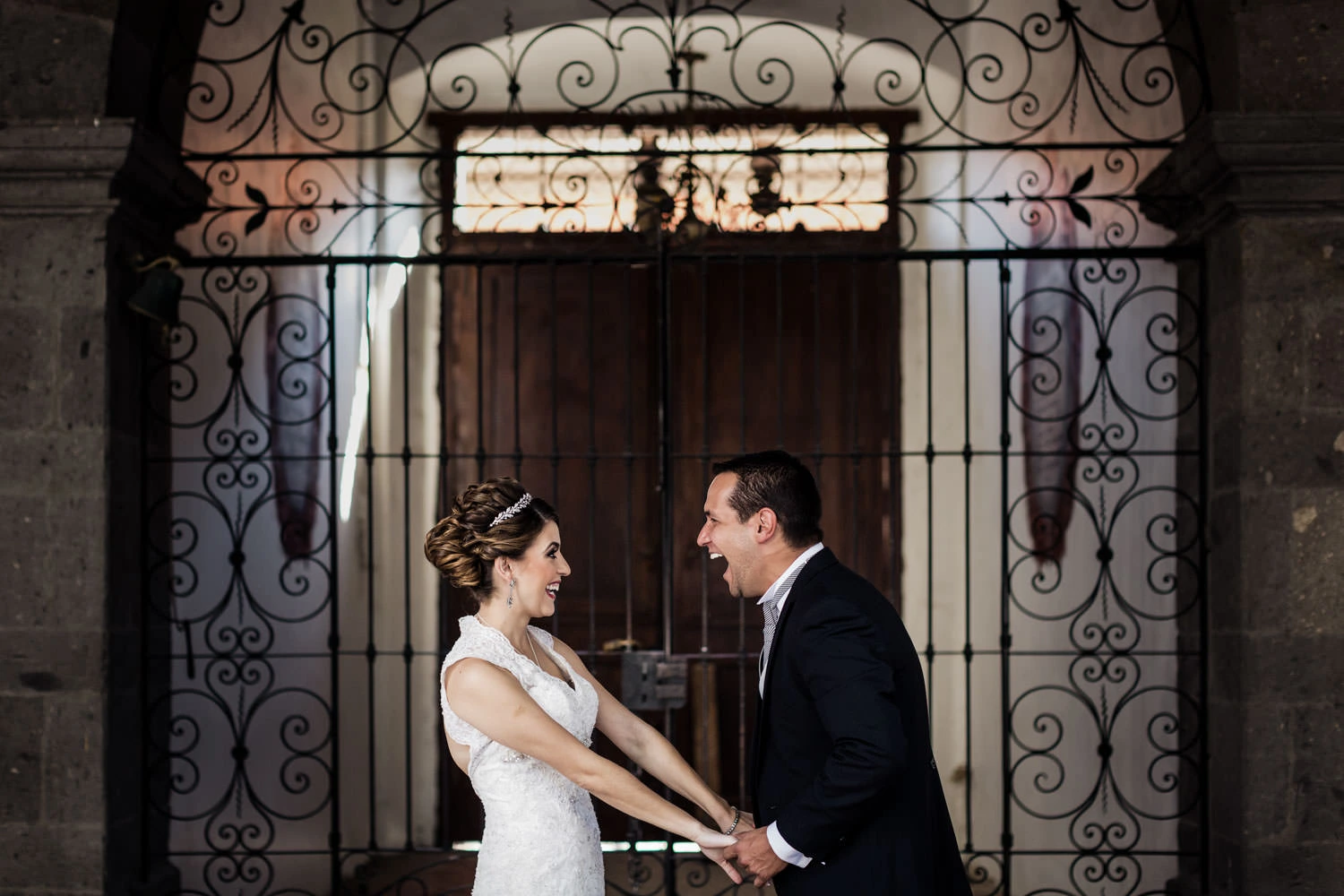 A bride and groom stand facing each other, holding hands and smiling joyfully. They are in front of an ornate wrought iron gate with stonework and warm lighting in the background. The bride wears a white dress, and the groom is in a black suit.