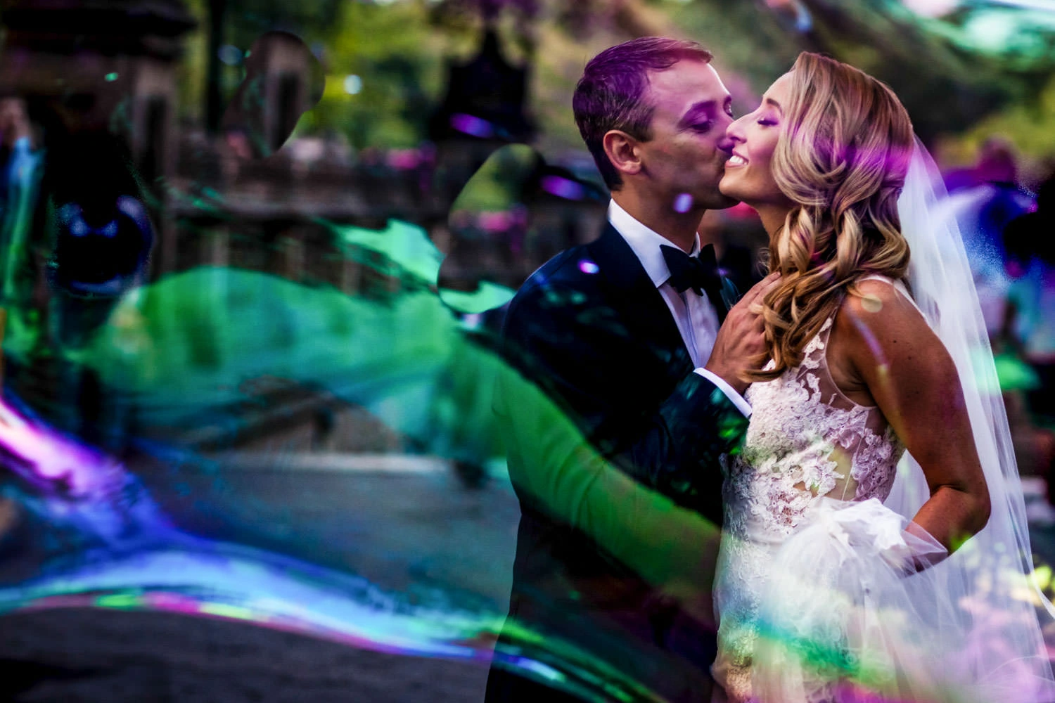 A bride and groom share a romantic moment, surrounded by colorful reflections. The groom in a tuxedo kisses the bride, who is in a floral lace gown and veil. They are enveloped in vibrant, swirled colors, suggesting a dreamy atmosphere.