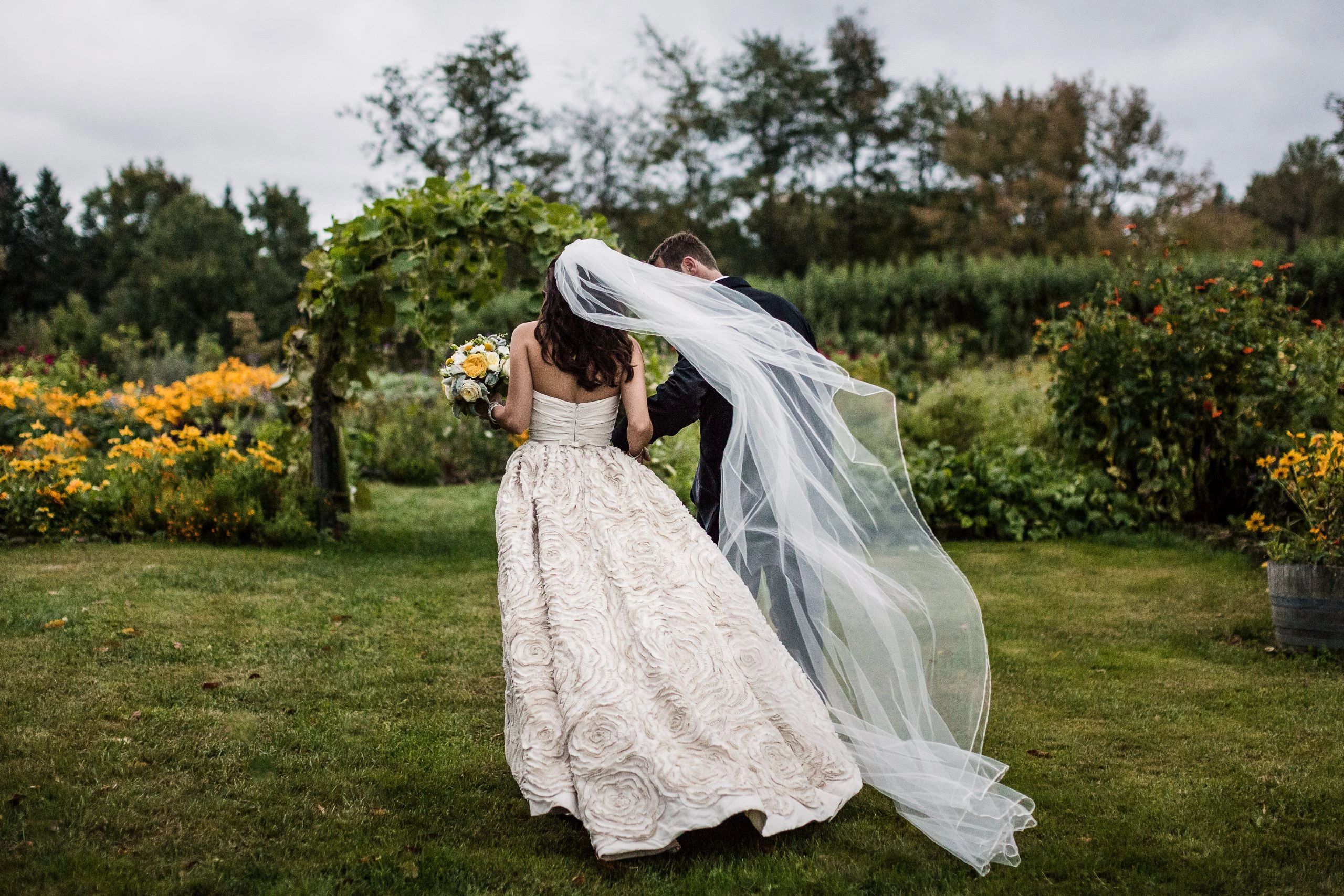 A bride and groom walk hand in hand through a garden. The bride wears a rose-patterned dress and a flowing veil, holding a bouquet. They are surrounded by lush greenery and vibrant flowers under an overcast sky.