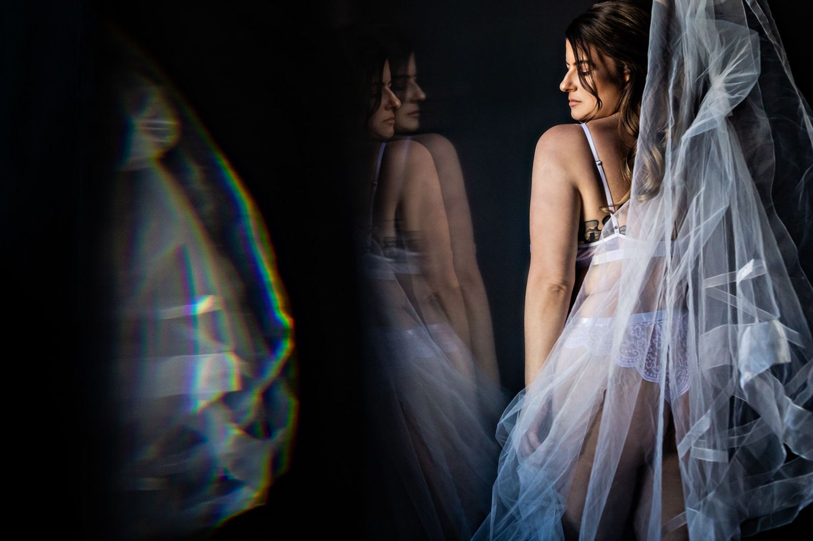 A woman stands in profile, wearing white lingerie with a sheer veil draped over her. Her reflection is visible in the background, alongside a prismatic light effect on the left. The scene is softly lit, emphasizing elegance and intimacy.