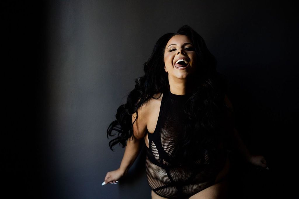 A woman with long dark hair laughs joyfully against a dark background. She is wearing a black mesh outfit, and her expression exudes confidence and happiness.