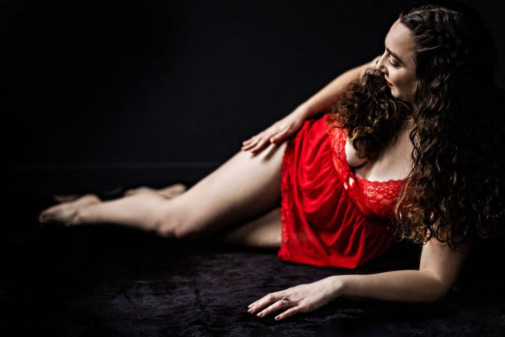 Woman lying on a dark surface, wearing a red lace outfit. Their long, curly hair cascades over one shoulder, and they are looking to the side with a gentle smile. The ambiance is intimate with soft lighting.