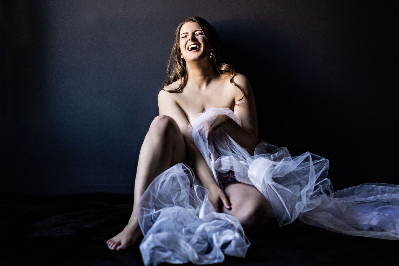 A woman with long hair sits against a dark background, draped in sheer white fabric. She is laughing, with her head tilted back, and her eyes closed. The image is artistically lit, emphasizing the textures of the fabric and her expression.