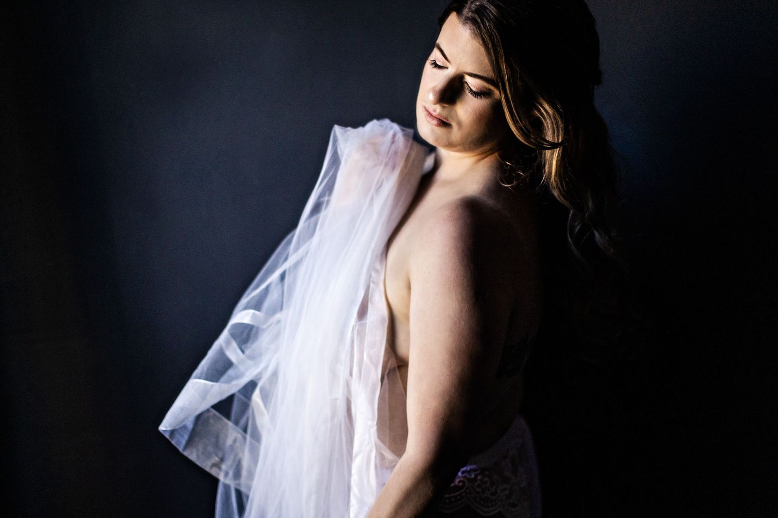 A woman with long hair poses with sheer fabric draped over her shoulder against a dark background. Her eyes are gently closed, and soft light highlights her face, creating a serene and contemplative mood.