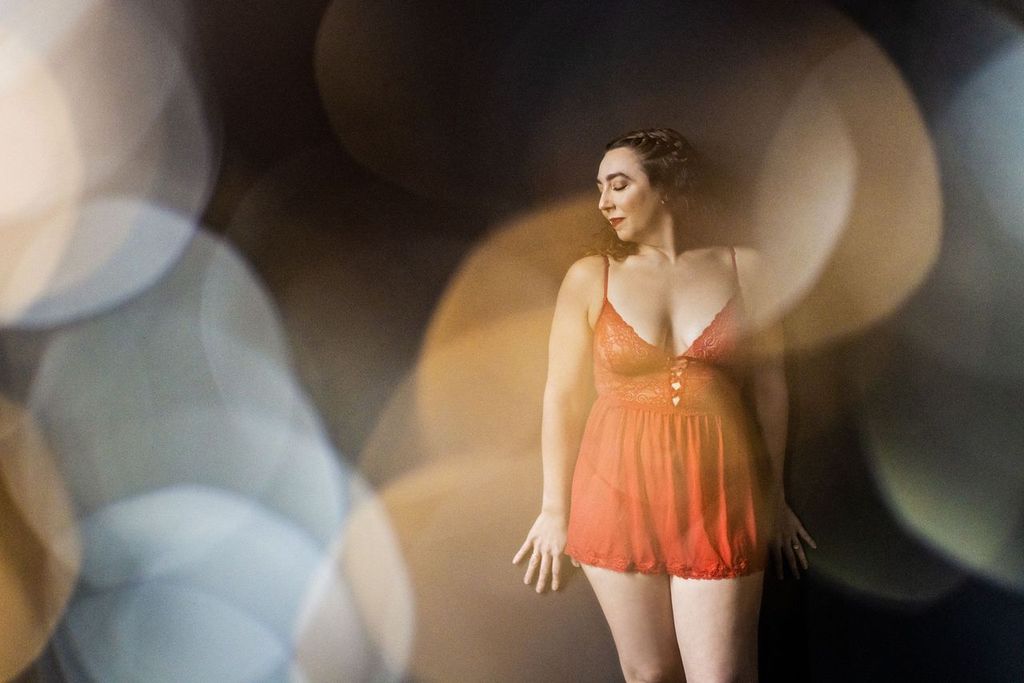 A woman stands confidently against a dark background, wearing a red, sheer, lace babydoll outfit. The foreground is filled with soft, out-of-focus bokeh lights, creating a dreamy effect. Her expression is serene, looking down to her side.