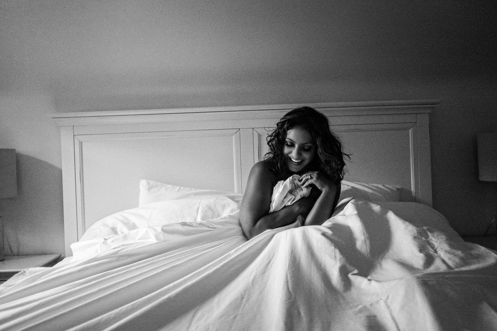 A woman sits on a bed, wrapped in a blanket, smiling warmly. The room is softly lit, creating a cozy and serene atmosphere. She is surrounded by white bedding and a headboard visible behind her.