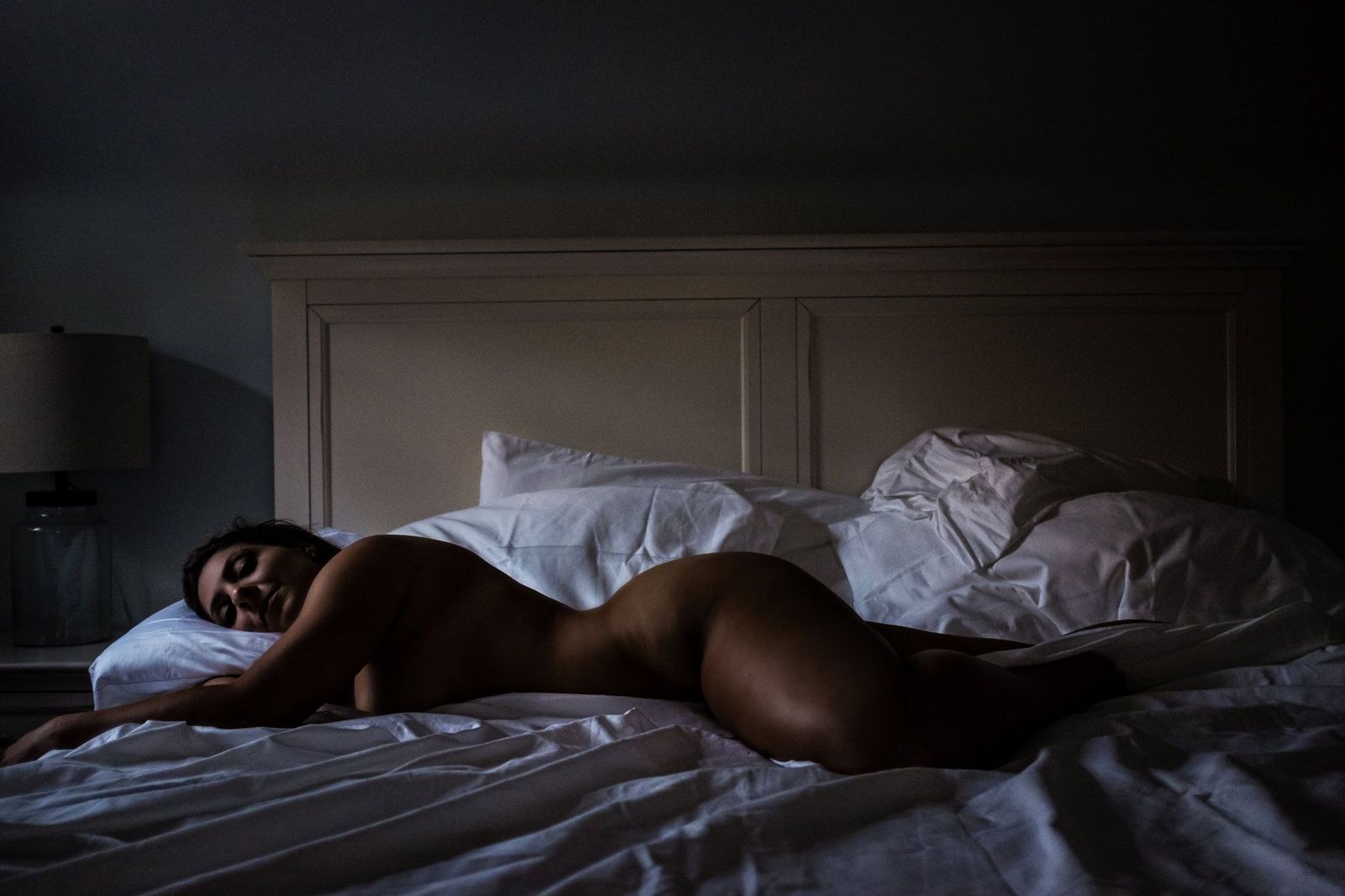 A woman lying on their side in bed, covered partially by a white sheet. The room is dimly lit, creating a serene and intimate atmosphere. A bedside lamp is visible in the background.