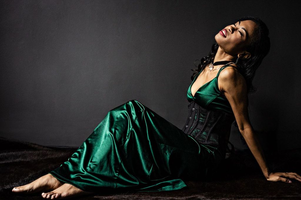A woman in a green satin gown and black corset leans back against a dark background, eyes closed, with a serene expression. She is barefoot, sitting on a textured surface, with her long hair cascading over her shoulders.