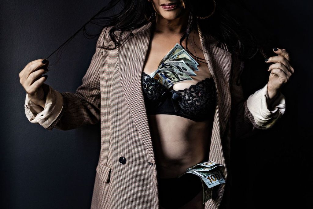 A woman wearing a suit jacket over lingerie with money tucked into the bra. The person is pulling on their hair, and a shadowy background contrasts with the scene.