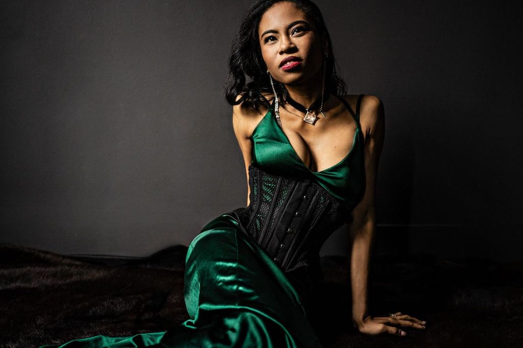 A woman in a long, green satin dress and black corset sits against a dark background, looking off to the side. Her hair is down, and she wears a necklace. The lighting creates a dramatic contrast.