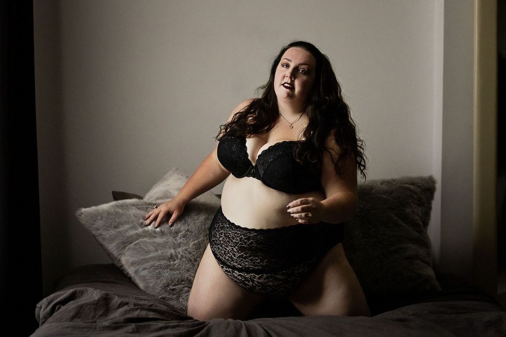 A woman with long, dark hair poses confidently on a bed. They are wearing a black lace bra and matching underwear. The setting is softly lit, with plush pillows and a neutral-colored background.