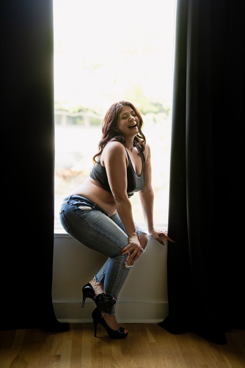 A woman wearing a black top, ripped jeans, and heels poses confidently in front of a large window with black curtains. She is smiling and has long, wavy hair. The room has wooden flooring and bright, natural light.