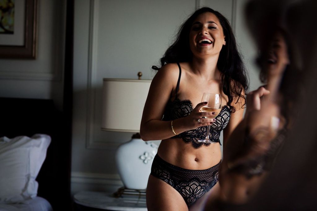 A woman in black lace lingerie stands by a bed, holding a glass of champagne. She is smiling joyfully, with soft lighting illuminating the elegant room, which features a lamp and framed artwork.