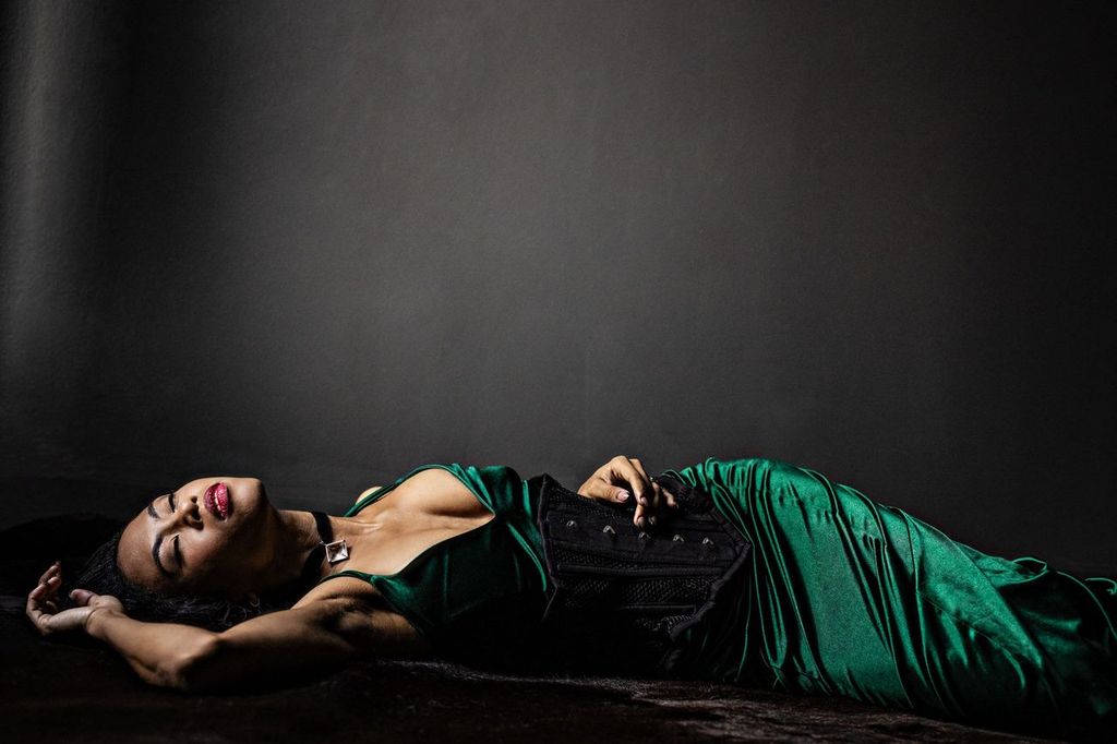 A woman in a green satin dress reclines on a dark surface against a gray backdrop. Their eyes are closed, and one arm rests above their head, creating a relaxed and serene pose.