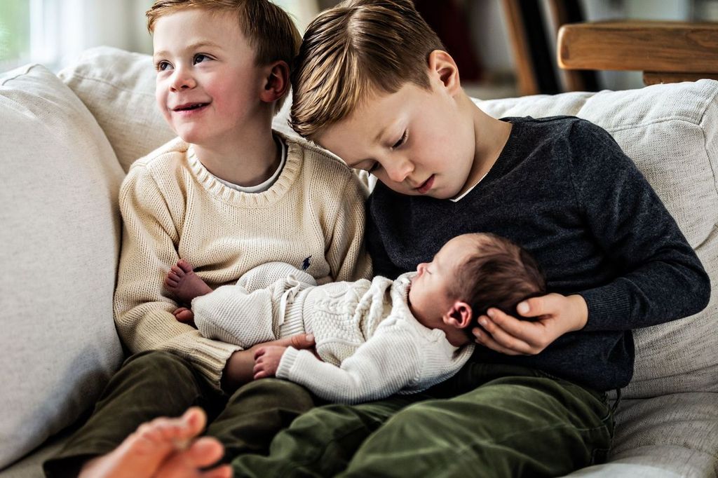 Two young boys are sitting on a couch. One is smiling and looking up, while the other gently holds a sleeping newborn in his lap. They are wearing cozy sweaters and sitting on a white sofa.
