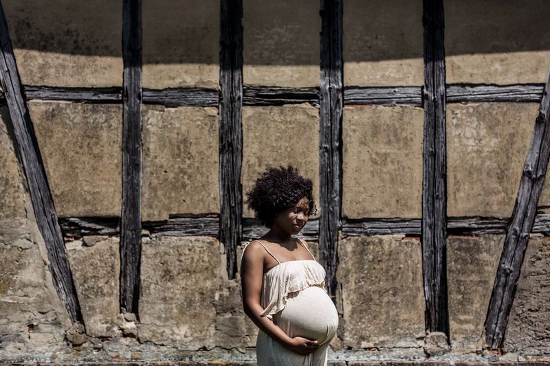 A pregnant woman with curly hair stands in a white dress, holding her belly. She is positioned in front of a rustic, wooden beam wall, casting a shadow in the sunlight.