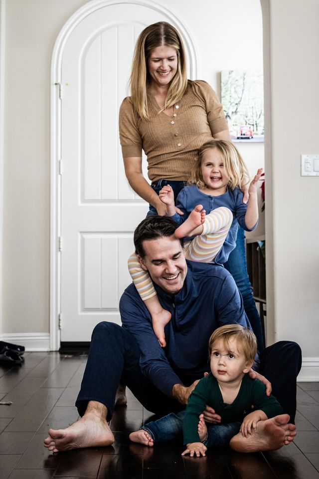 A smiling family is playfully gathered in a hallway. A mother stands while a father crouches, holding a laughing toddler on his shoulders. Another child, seated on the floor, looks curious. The scene is light-hearted and joyful.