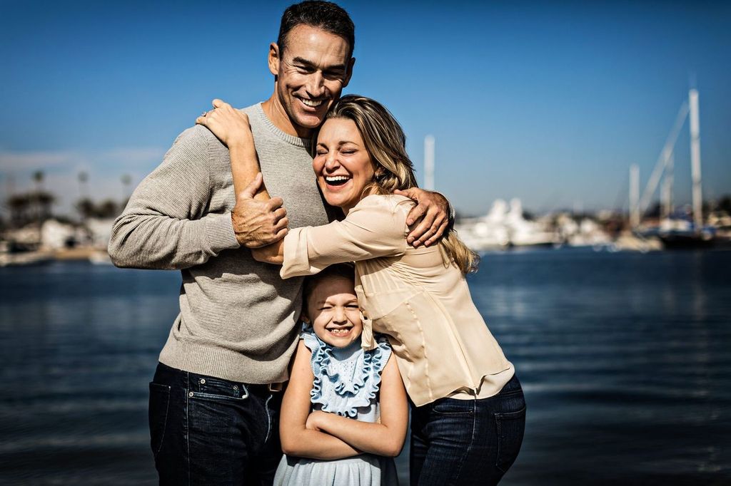 A joyful family moment by the water: an adult couple and a young girl embrace and smile broadly against a backdrop of boats and a clear blue sky. The adults are wearing casual sweaters, and the girl is in a blue dress.