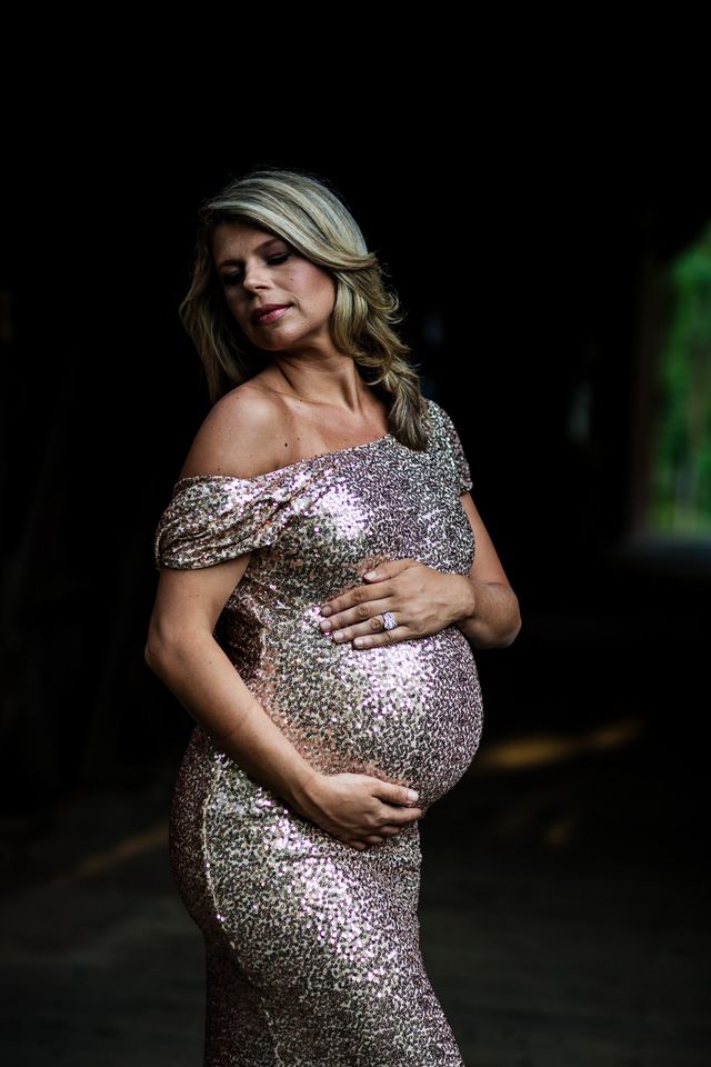 A pregnant woman in a sparkling off-shoulder dress gently cradles her belly. She stands against a dark background, her eyes closed and expression serene. The scene conveys a sense of calm and anticipation.