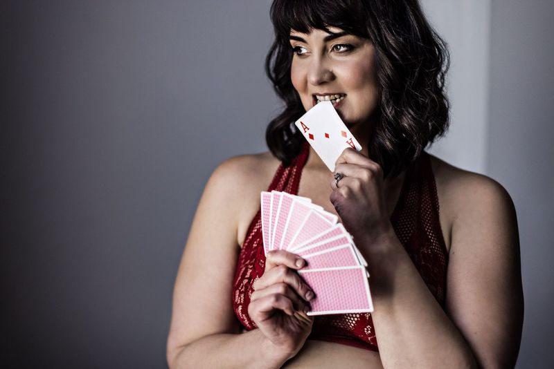 A woman in a red outfit holds a fan of playing cards in one hand and bites an ace of hearts with a playful expression. The background is a neutral gray.