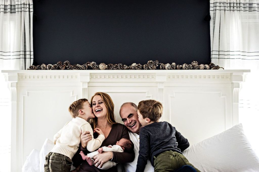 A family is sitting on a bed with the parents in the center holding a baby. Two young boys are playfully leaning on either side of them. The background features a white mantel with pinecones on top. The atmosphere is joyful and relaxed.