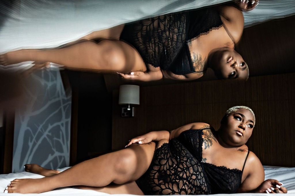 A person wearing black lace lingerie is lying on a bed. Their reflection is visible in a mirrored surface above. The background features a lit table lamp and abstract wall patterns, creating a moody ambiance.