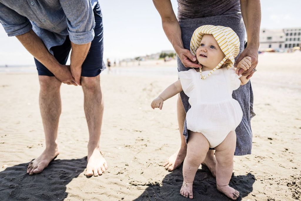 A baby wearing a white outfit and sun hat walks on a sandy beach, supported by an adult holding their hands. Another adult stands nearby, bent slightly forward. The beach is expansive, with buildings in the distance under a clear sky.