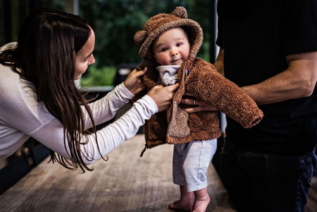 A smiling baby stands on a wooden table, wearing a brown fuzzy jacket with bear ears. An adult woman adjusts the jacket while another adult supports the baby. The background is blurred, suggesting a cozy indoor setting.