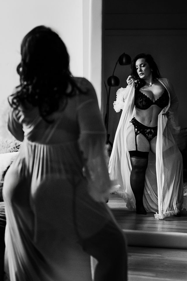 A woman in a sheer robe, lingerie, and stockings stands confidently in front of a mirror, her reflection visible. The scene, captured in black and white, conveys a sense of empowerment and self-assurance.