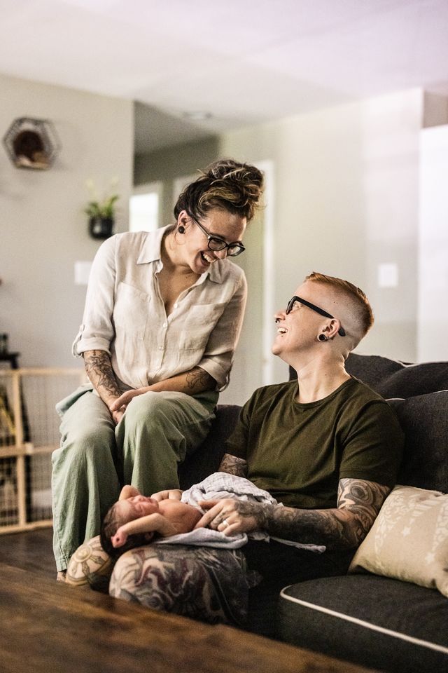 A smiling couple sits in a living room with a baby. One person, with short hair and glasses, holds the baby on their lap. The other, with long hair, leans over, smiling down. The room is softly lit and cozy.