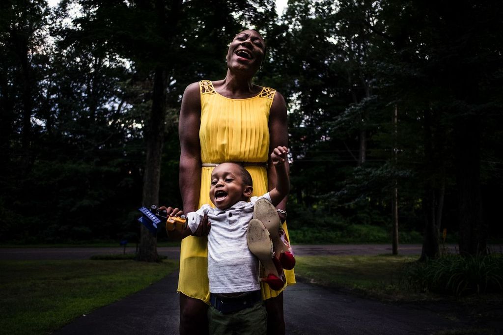 A joyful woman in a yellow dress stands on a dimly lit path surrounded by trees, holding a smiling child who is lifting his arms. The child is holding a stuffed toy and a book. Both appear to be laughing.