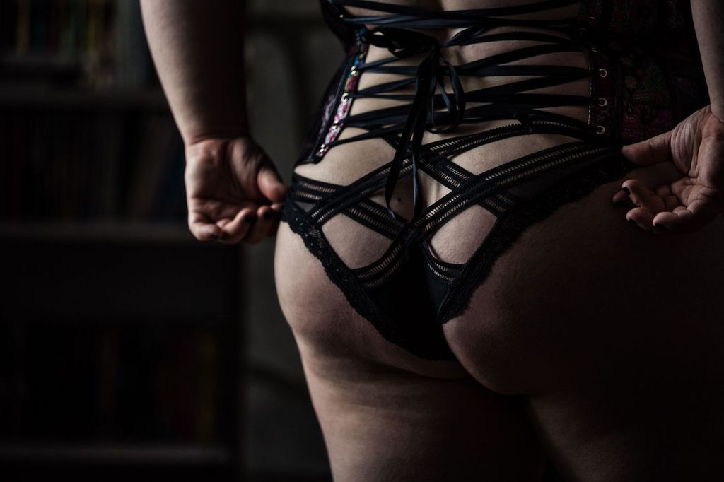 Person wearing black lingerie with lace and strap details, seen from behind in a dimly lit room.