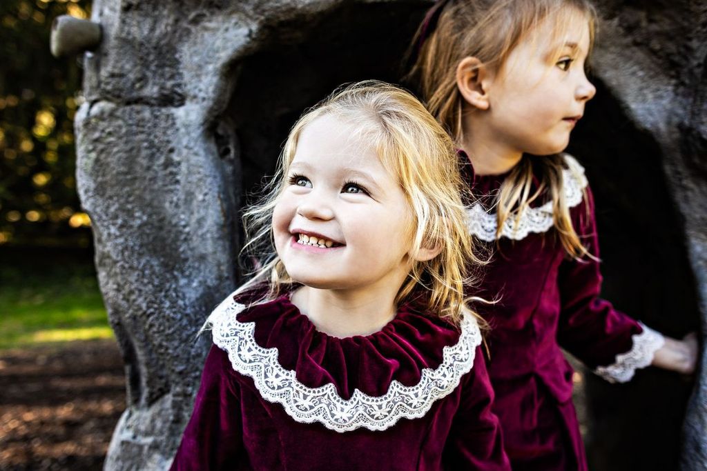Two young girls in matching velvet dresses with white lace collars. One smiles while looking up, and the other gazes to the side. They stand in front of a rock-like structure in an outdoor setting with soft natural lighting.