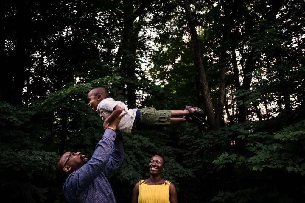 A man joyfully lifts a smiling child into the air in a forest. A woman stands nearby, smiling and watching them. The scene is surrounded by lush green trees, and the lighting suggests its either early evening or late afternoon.