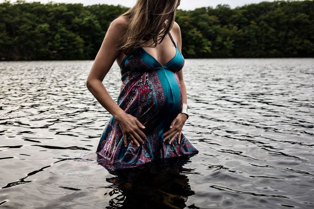 Pregnant woman stands in a lake with water up to her thighs. She wears a colorful dress and holds her belly, surrounded by a forested shoreline. The scene is serene, with soft lighting and a peaceful atmosphere.