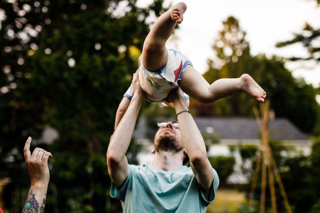 An adult in a teal shirt playfully lifts a baby wearing a white outfit with colorful stars overhead. Another arm is raised beside them. The background features trees and blurred houses, suggesting a backyard setting.