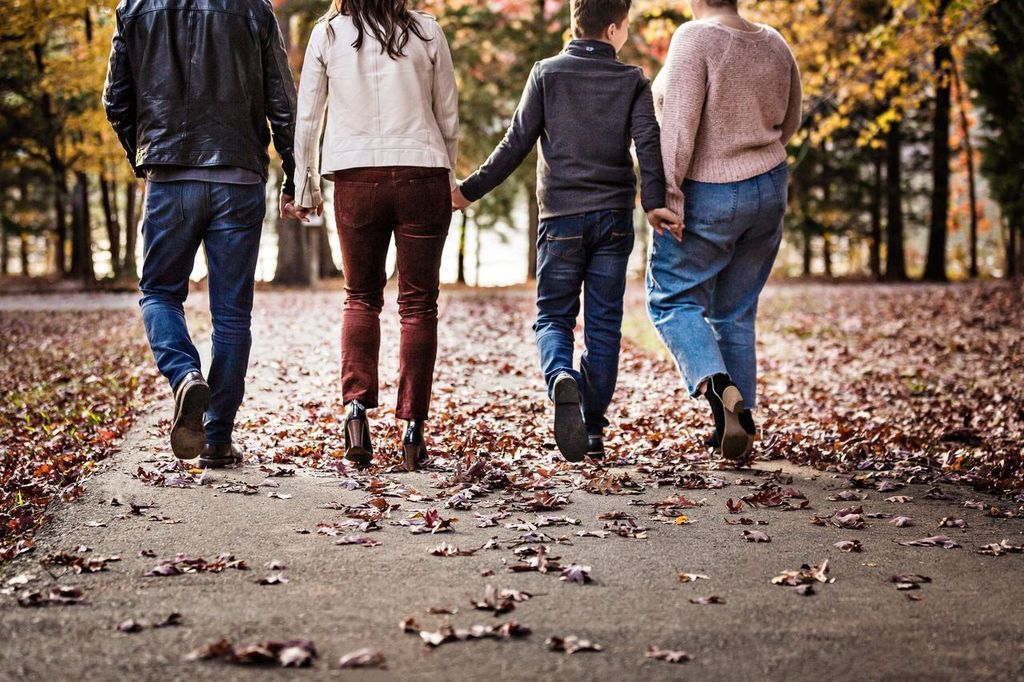 Four people are walking on a leaf-covered path in a forested area during autumn. They are holding hands in pairs, dressed in casual fall clothing, with leaves scattered around. The trees have colorful foliage in the background.