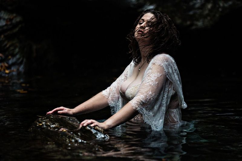A woman in a sheer, white lace shawl stands partly submerged in dark water. Her hair is wet, and she leans back on a rock, appearing relaxed and serene. The background is shadowy and natural.
