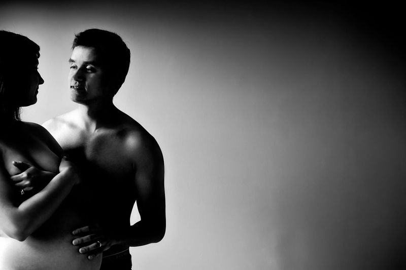 A black and white photo of a shirtless man and a pregnant woman standing close together. The woman cradles her belly while gazing at the man, who looks at her. The background is a soft gradient from light to dark on the right side.