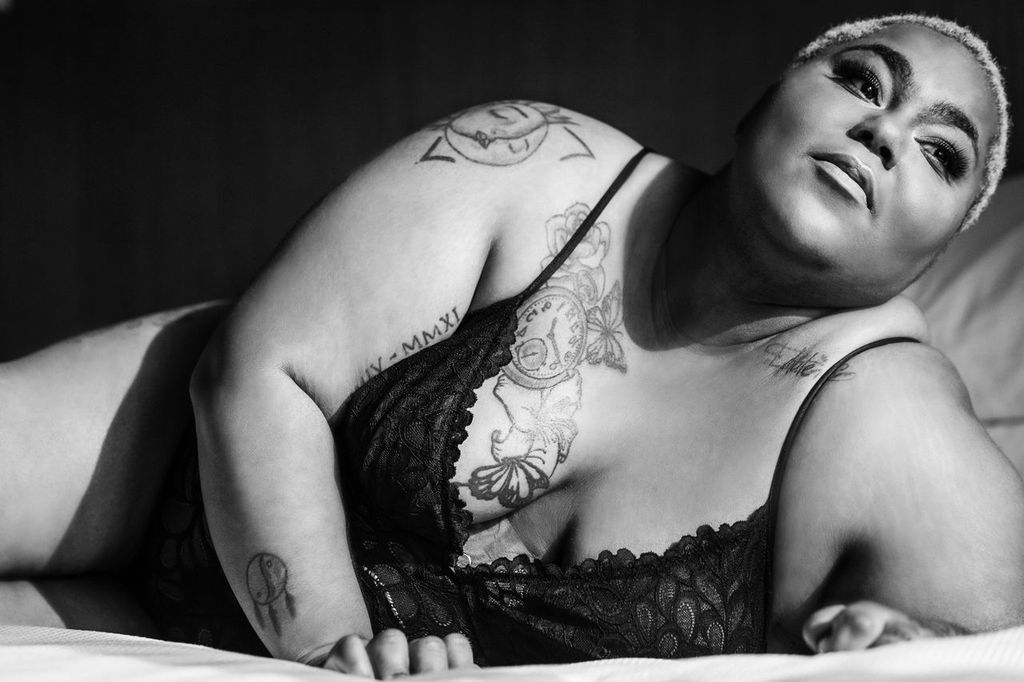 Black and white photo of a woman with short hair lying on a bed. She is wearing lace clothing and have tattoos on their arms and shoulder. The image captures a serene and confident expression.