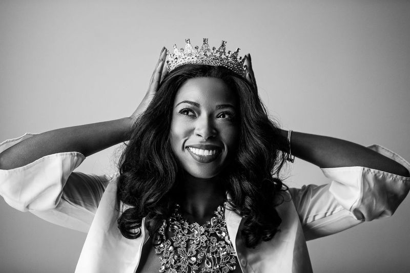 A black and white photo of a woman smiling broadly while adjusting a crown on her head. She has long, wavy hair and are wearing an ornate, embellished top under a blazer.
