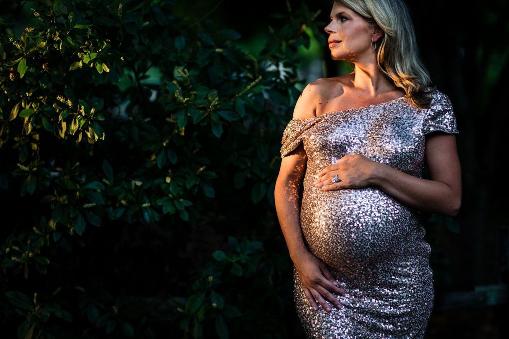A pregnant woman in a sparkling, off-shoulder dress stands under dappled sunlight. She gently cradles her belly with one hand, surrounded by dark green foliage. Her expression is serene and thoughtful.