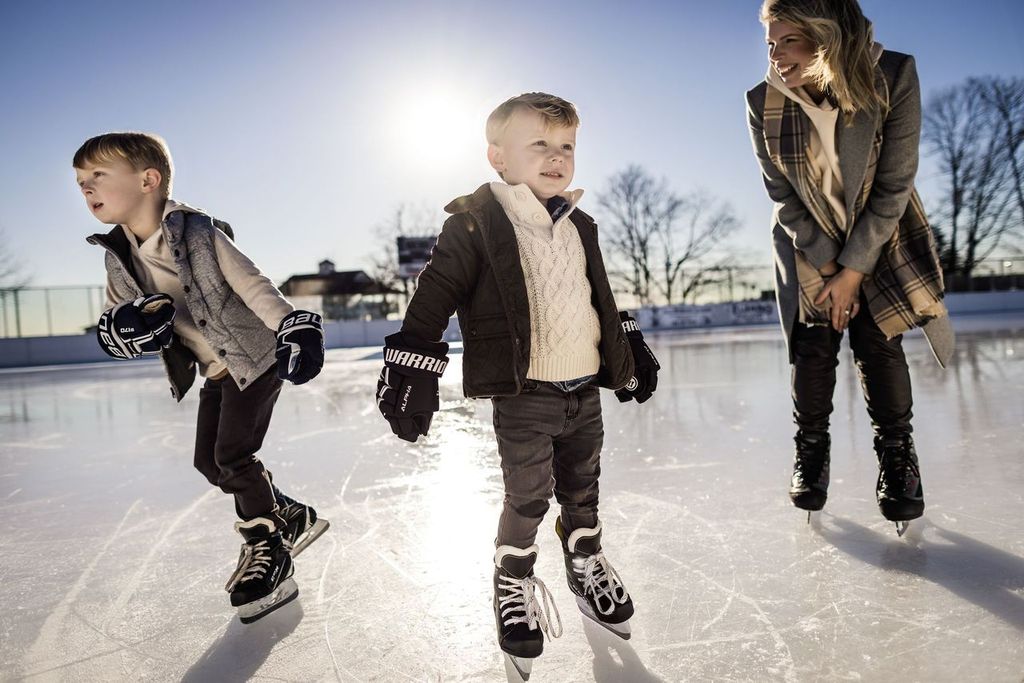 A woman and two young children ice skating outdoors on a sunny day. The children are wearing warm jackets and gloves, while the woman is wearing a scarf. They all appear to be enjoying the activity.