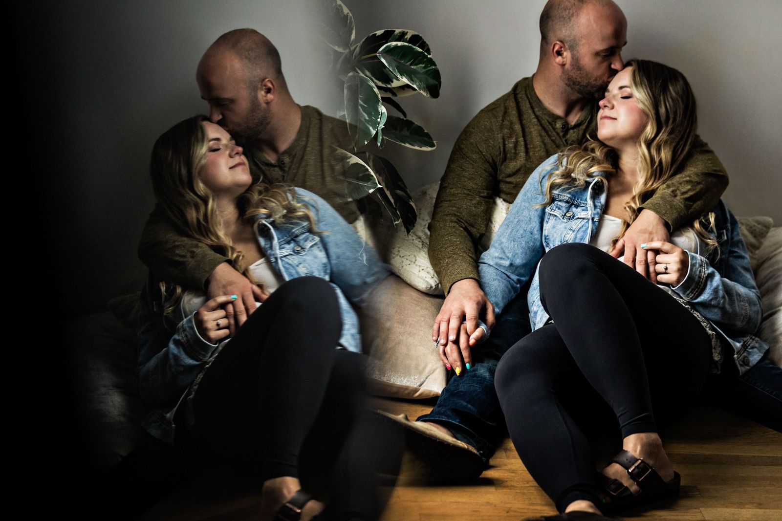 A couple sits on the floor, embracing lovingly. The man kisses the womans forehead as they hold hands. Theyre both in casual clothing, with a plant in the background. A soft reflection of them is visible on the left.