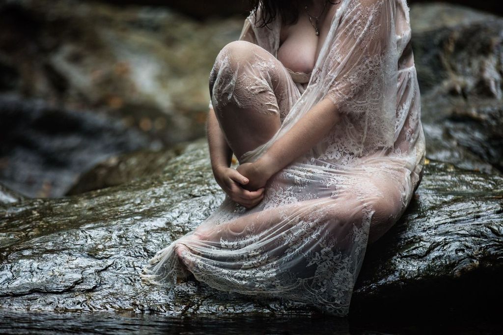 Person sitting on a rock by a stream, draped in a translucent, lacy shawl. The scene is natural and serene, with soft lighting highlighting the delicate fabric.